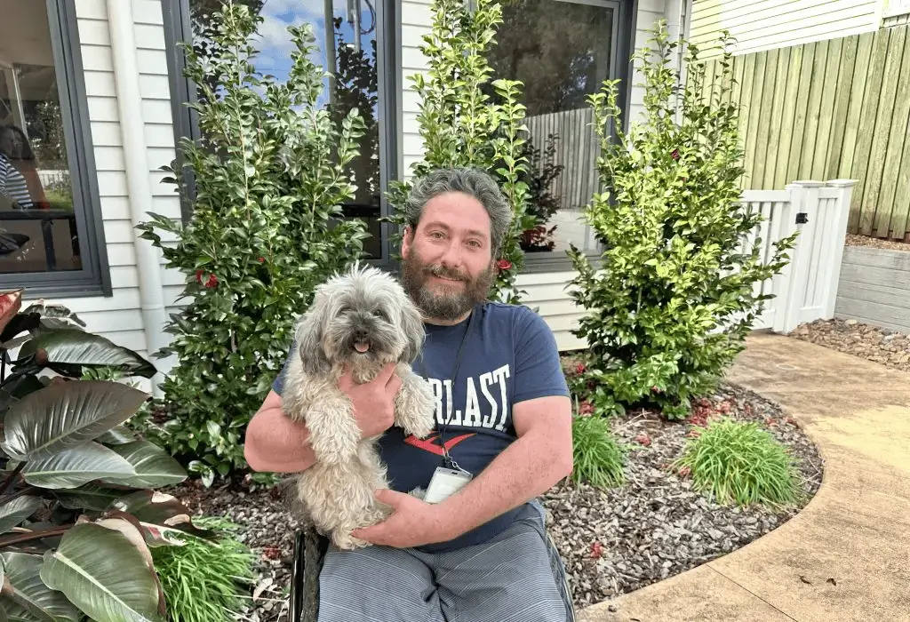 Man in a wheelchair holding a dog outdoors and smiling.