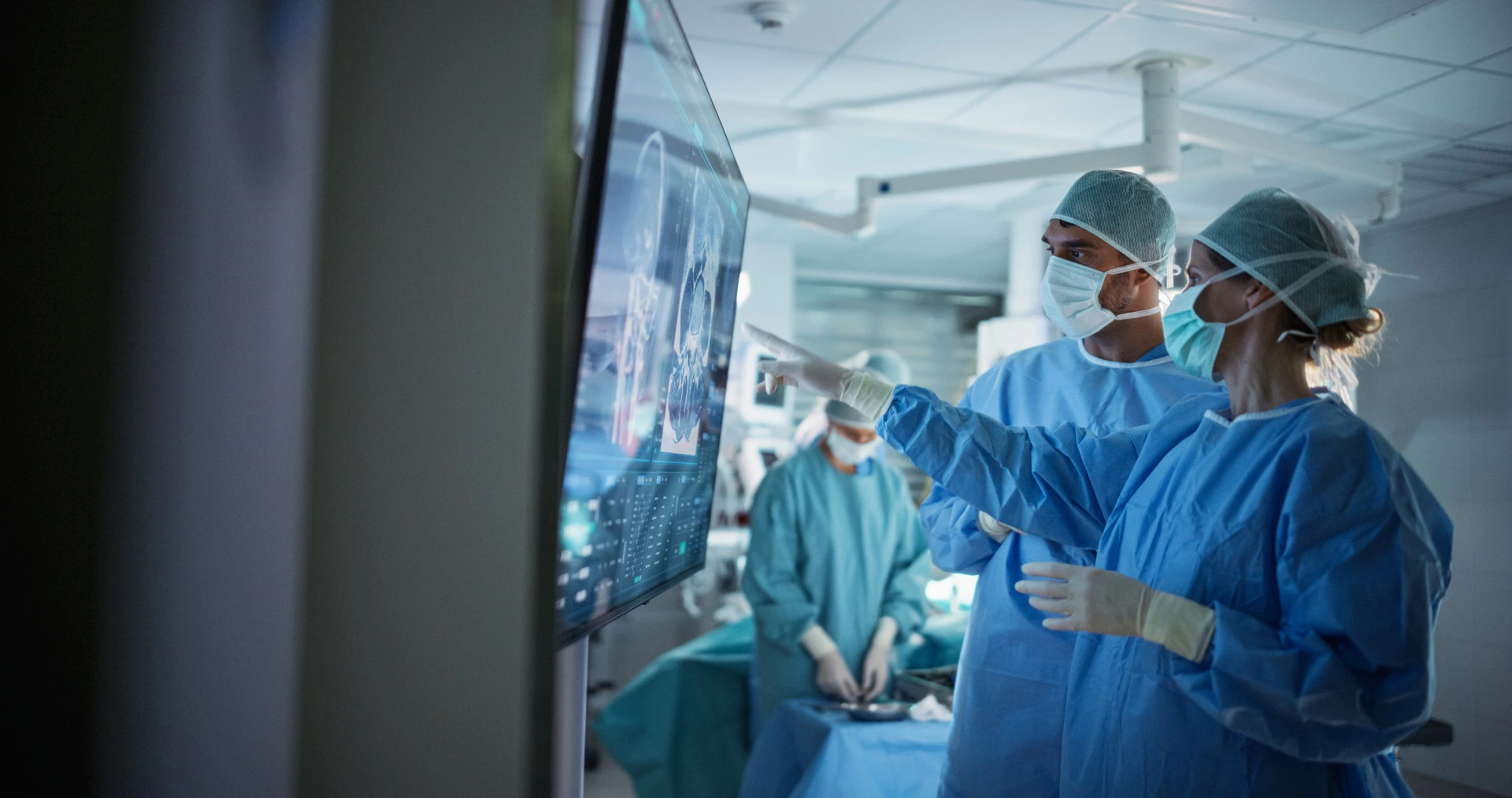 Doctors in surgical gowns and masks review images on a large digital screen in an operating room while other medical staff work in the background, preparing for or performing surgery.