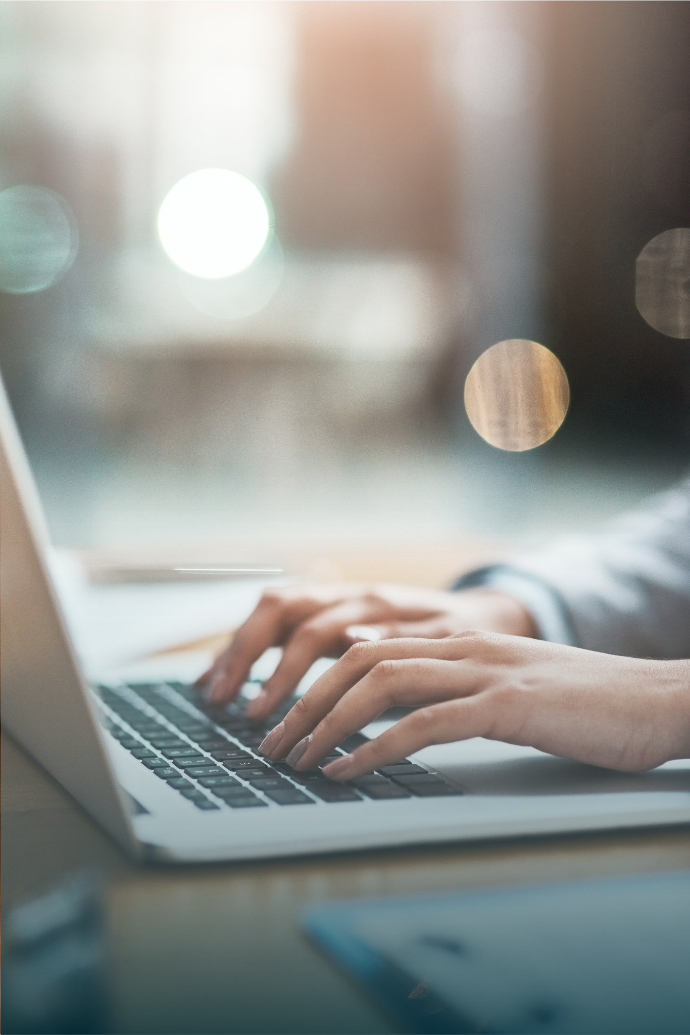A person’s hands typing on a laptop keyboard in a softly lit setting, with blurred lights and documents in the background.