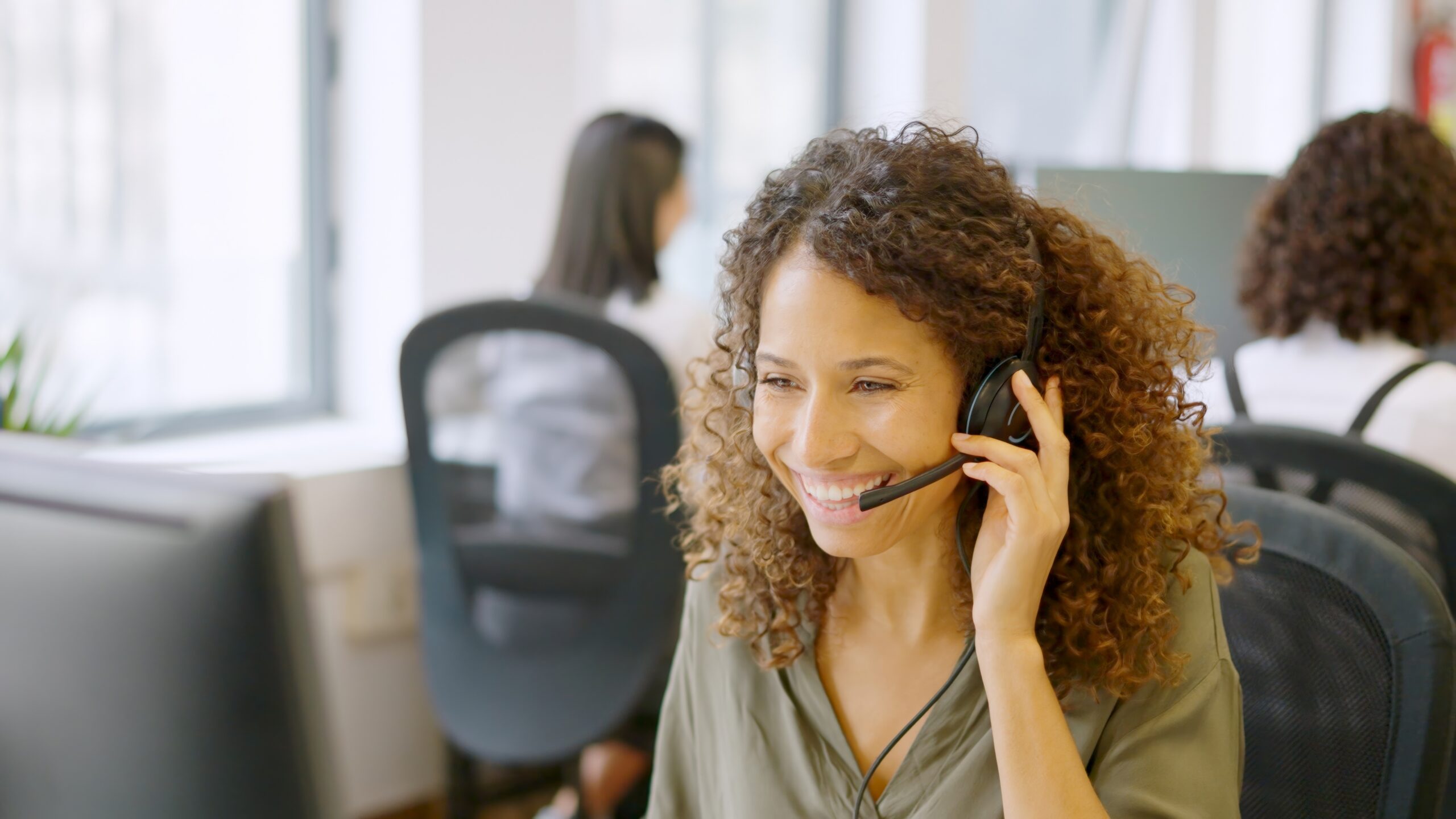 A smiling woman with curly hair wearing a headset sits at a desk in a bright office, talking on the phone. Another person is working at a computer in the background.