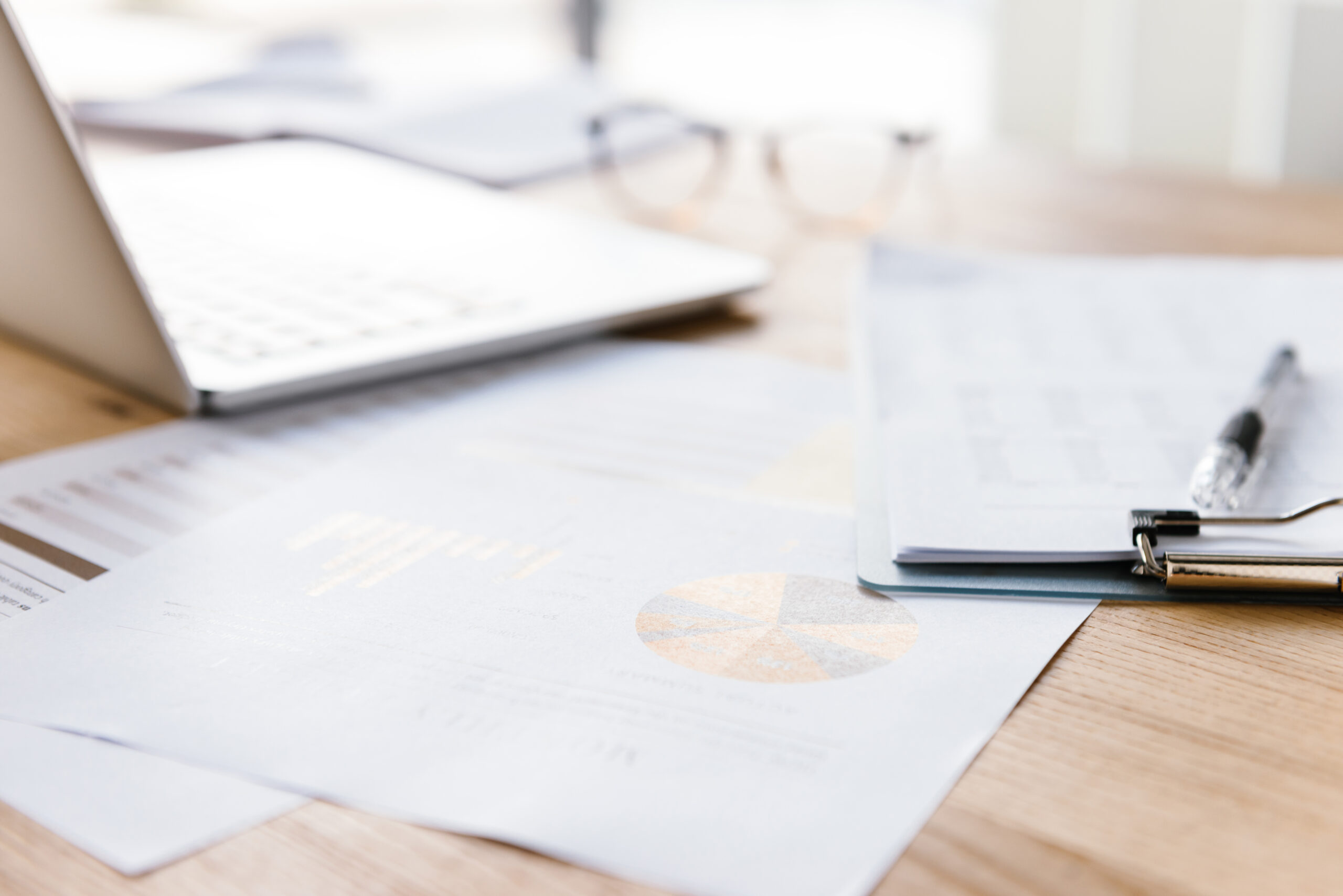 A close-up of a desk with financial documents, a pen, an open laptop, and a pair of eyeglasses, suggesting a work or study environment. The papers display charts and graphs.