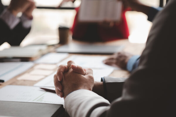 Close-up of a person’s hands clasped on a table during a meeting, with documents and blurred people in the background.